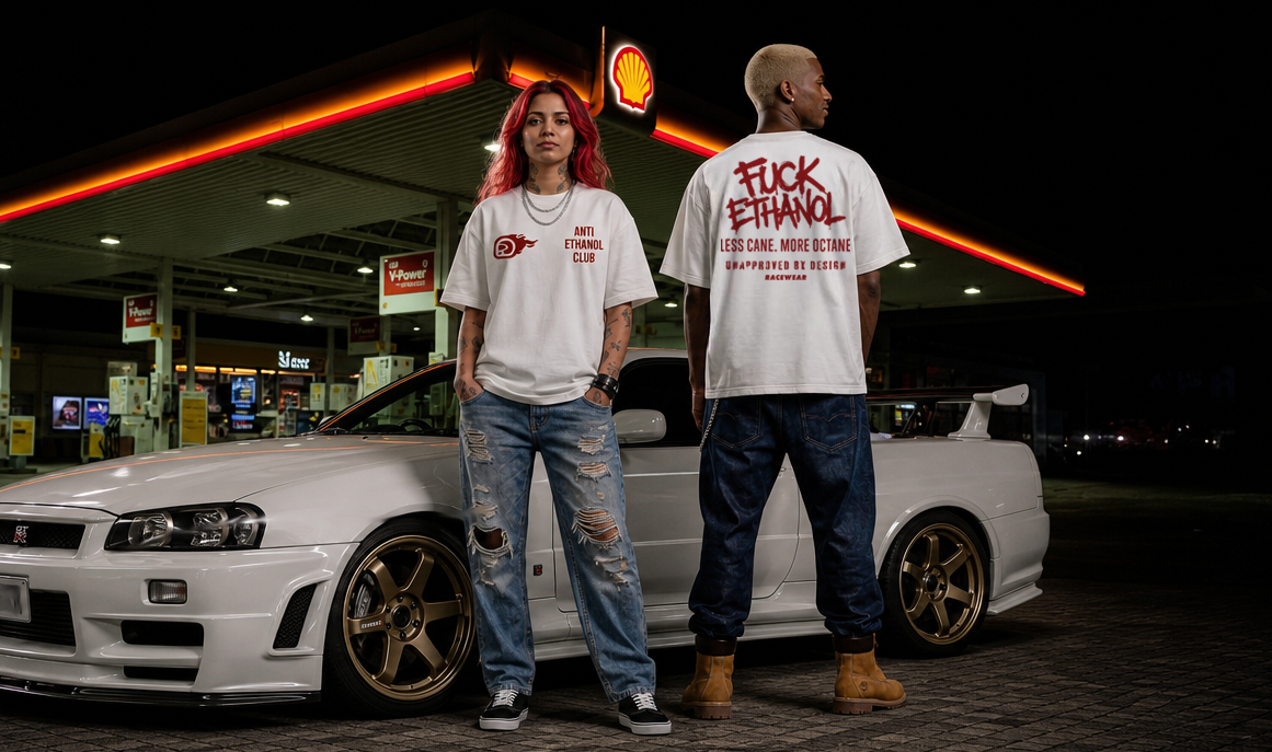 Two models wearing white graphic t-shirts from Racewear India stand next to a white modified Nissan R33 sports car at a gas station at night. The model on the left has red hair and wears distressed blue jeans, while the model on the right has a shaved head and wears dark jeans and brown boots.
